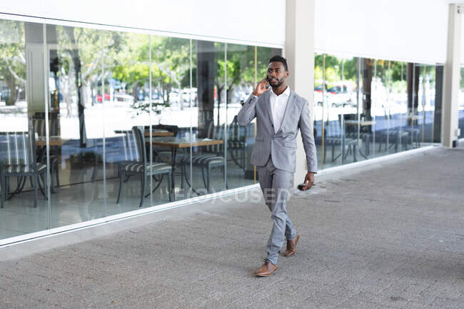 Homem de negócios afro-americano andando usando máscara facial falando no smartphone. homem de negócios em movimento na cidade. — Fotografia de Stock