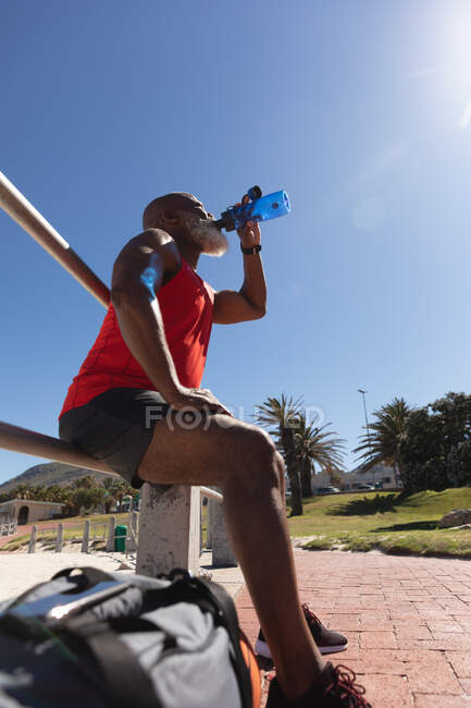 Fit senior homme afro-américain exercice assis au soleil boire de la bouteille d'eau. retraite saine mode de vie en plein air fitness. — Photo de stock