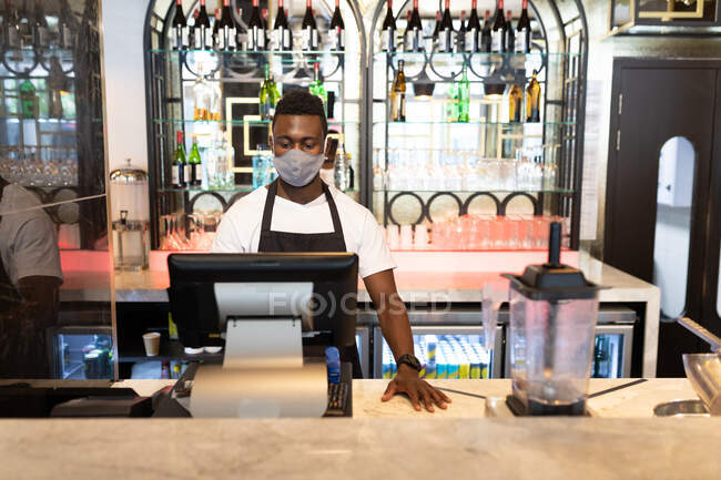 African american male barista wearing face mask using cash register. health and hygiene in business during coronavirus covid 19 pandemic. — Stock Photo