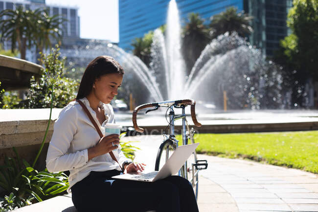 Femme afro-américaine avec tasse de café à l'aide d'un ordinateur portable tout en étant assis au parc d'entreprise. mode de vie concept de vie pendant coronavirus covid 19 pandémie. — Photo de stock