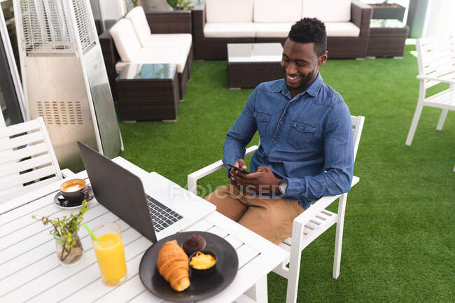 Hombre afroamericano sentado en un café con smartphone y portátil desayunando. hombre de negocios en la salida en la ciudad. - foto de stock