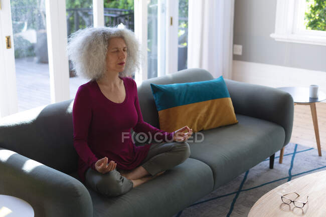 Senior caucasian woman sitting on couch in living room meditating. staying at home in self isolation during quarantine lockdown. — Stock Photo