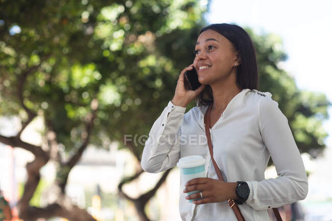 Африканская американка с кофейной чашкой разговаривает по смартфону на улице. Концепция образа жизни во время пандемии коронавируса 19. — стоковое фото