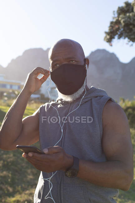 Fit senior african american man wearing face mask putting on earphones. healthy retirement technology communication outdoor fitness lifestyle hygiene during coronavirus covid 19 pandemic. — Stock Photo