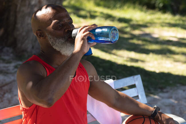 Ajuste o homem americano africano sênior que está sentado no parque que bebe da garrafa de água. esporte de aposentadoria saudável ao ar livre fitness lifestyle. — Fotografia de Stock