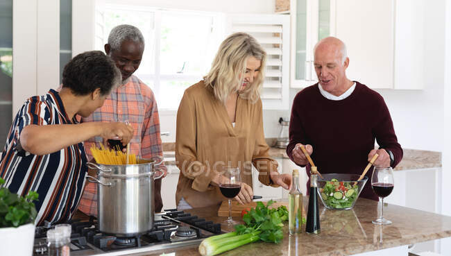 Senior caucasian and african american couples making pasta and salad at home. senior retirement lifestyle friends socializing. — Stock Photo