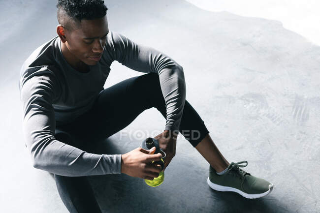 African american man sitting in empty urban building and resting after playing basketball. holding a bottle of water. urban fitness healthy lifestyle. — Stock Photo