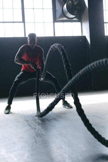 Homme afro-américain portant des vêtements de sport luttant contre les cordes dans un bâtiment urbain vide. forme physique urbaine mode de vie sain. — Photo de stock