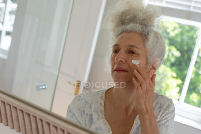 Старша жінка-какажан стоїть у ванній і кладе вершки на обличчя. Перебуваючи вдома в ізоляції під час карантину.. — стокове фото