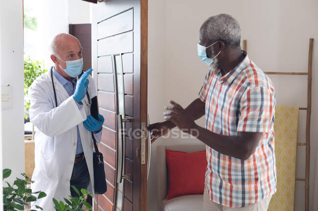 Senior african american man greeting caucasian senior doctor both wearing masks at home. healthcare hygiene protection during coronavirus covid 19 pandemic. — Stock Photo
