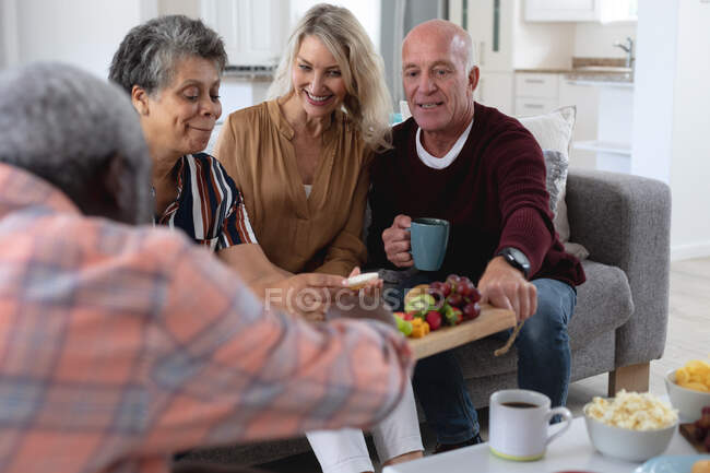Casais seniores caucasianos e afro-americanos sentados à mesa comendo queijo e frutas em casa. sênior aposentadoria estilo de vida amigos socialização. — Fotografia de Stock