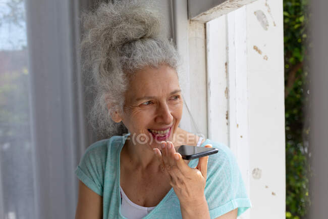 Femme caucasienne âgée debout par la fenêtre parlant sur smartphone à la maison. rester à la maison en isolement personnel pendant le confinement en quarantaine. — Photo de stock