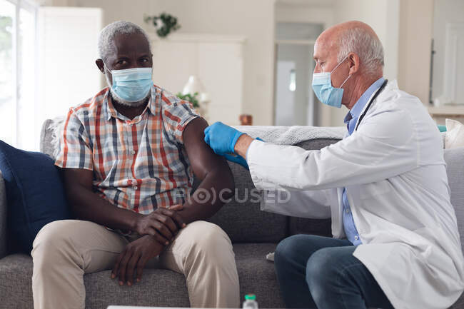 Senior caucasian male doctor vaccinating male patient both wearing face masks at home. healthcare hygiene protection during coronavirus covid 19 pandemic. — Stock Photo