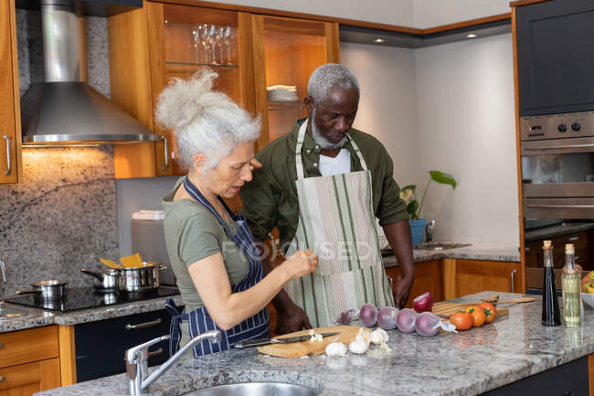Ein älteres gemischtes Paar steht in der Küche und schneidet Gemüse. Während der Quarantäne zu Hause bleiben und sich selbst isolieren. — Stockfoto