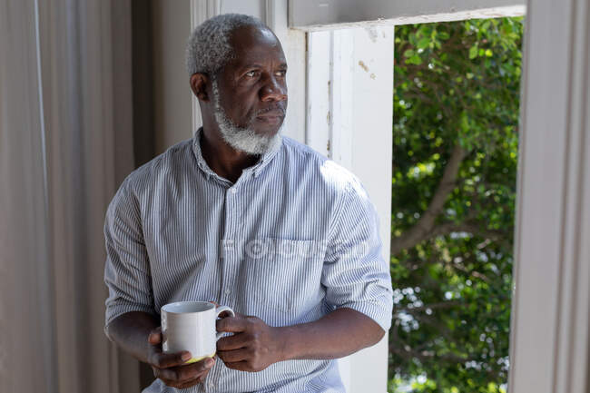 Senior african american man standing by window drinking coffee at home. staying at home in self isolation during quarantine lockdown. — Stock Photo