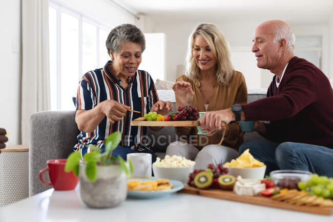 Couples supérieurs caucasiens et afro-américains assis à table mangeant du fromage et des fruits à la maison. senior retraite style de vie amis socialisation. — Photo de stock