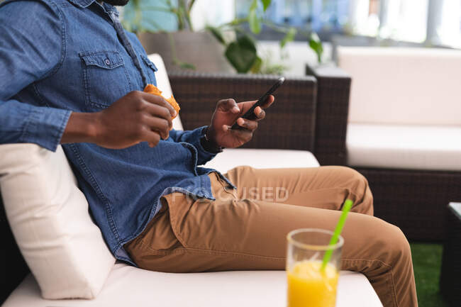 Afrikanischer Mann sitzt mit Smartphone in einem Café und isst Croissant. Geschäftsmann unterwegs in der Stadt. — Stockfoto