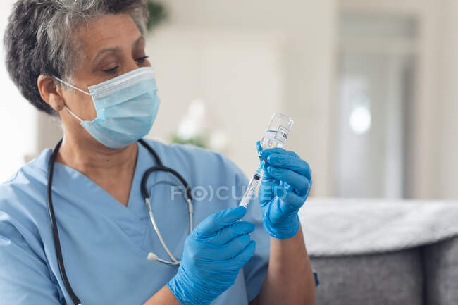 Senior african american female doctor wearing face mask preparing vaccination at home. healthcare hygiene protection during coronavirus covid 19 pandemic. — Stock Photo