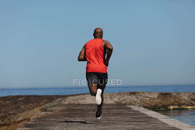 Visão traseira do homem americano africano sênior apto correndo no caminho costeiro. aposentadoria saudável ao ar livre fitness lifestyle. — Fotografia de Stock