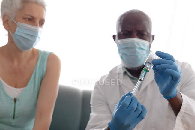 Medico anziano afroamericano di sesso maschile che prepara la vaccinazione covid 19 per anziana donna caucasica a casa. assistenza sanitaria e igiene a domicilio in isolamento personale durante l'isolamento in quarantena. — Foto stock