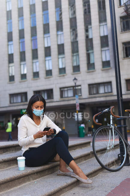 Femme afro-américaine portant un masque facial à l'aide d'un smartphone assis dans les escaliers à l'extérieur. mode de vie concept de vie pendant coronavirus covid 19 pandémie. — Photo de stock