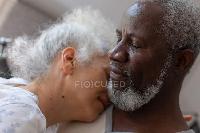 Senior pareja de raza mixta en el dormitorio sentado en la cama abrazando. permanecer en casa en aislamiento durante el bloqueo de cuarentena. - foto de stock