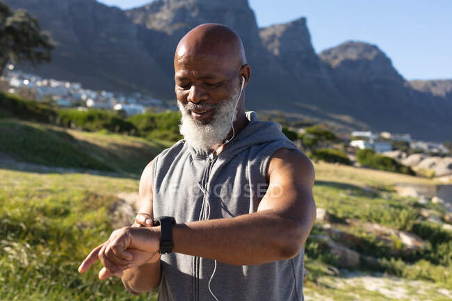 Fit senior african american man exercising wearing earphones checking smartwatch. healthy retirement technology communication outdoor fitness lifestyle. — Stock Photo