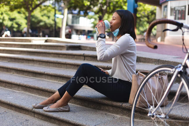 Mujer afroamericana con mascarilla baja bebiendo café mientras está sentada en las escaleras al aire libre. estilo de vida concepto de vida durante el coronavirus covid 19 pandemia. - foto de stock