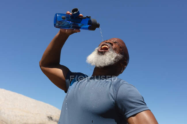 Ajustez l'homme afro-américain aîné exerçant la boisson de la bouteille d'eau contre le ciel bleu. retraite saine mode de vie en plein air fitness. — Photo de stock