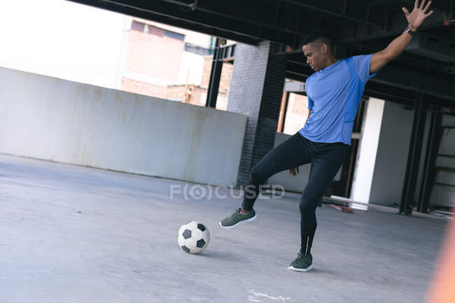 Hombre afroamericano haciendo trucos con un balón de fútbol en un edificio urbano vacío. aptitud urbana estilo de vida saludable. - foto de stock