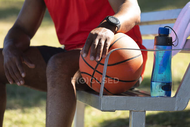 Sección media del hombre afroamericano en forma sentado en el parque con baloncesto y botella de agua. deporte de jubilación saludable estilo de vida fitness al aire libre. - foto de stock