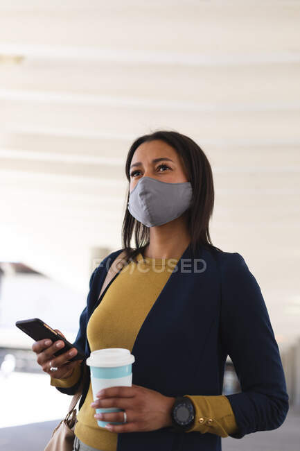 Donna afroamericana con maschera facciale che tiene in mano tazza di caffè e smartphone per strada. stile di vita durante il coronavirus covid 19 pandemia. — Foto stock