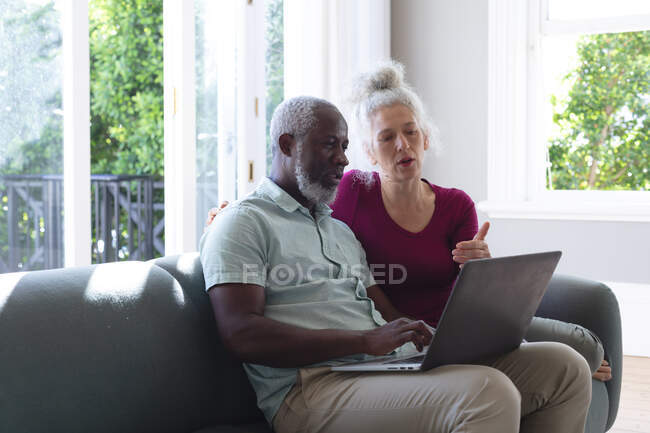 Couple mixte senior assis sur le canapé regardant ordinateur portable ensemble dans le salon. rester à la maison en isolement personnel pendant le confinement en quarantaine. — Photo de stock