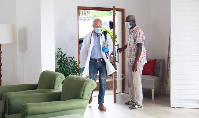 Старший афроамериканський чоловік вітає кавказького старшого лікаря, обидва носять маски вдома. Захист гігієни охорони здоров'я під час пандемії коронавірусу 19 . — стокове фото