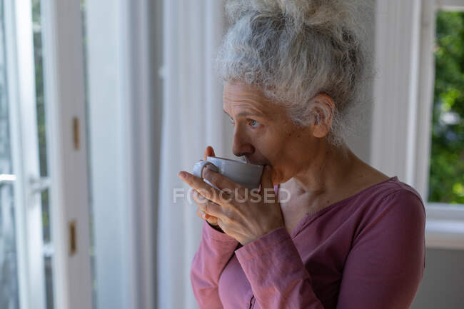 Старша жінка-кавоварка стоїть біля вікна і п'є каву вдома. Перебуваючи вдома в ізоляції під час карантину.. — стокове фото