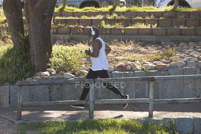 Adatto all'uomo anziano afroamericano che indossa cuffie wireless in esecuzione nel parco. sano stile di vita fitness all'aperto pensionamento. — Foto stock