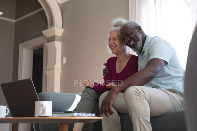 Sénior casal misto sentado no sofá abraçando usando laptop na sala de estar. ficar em casa em auto-isolamento durante o confinamento de quarentena. — Fotografia de Stock