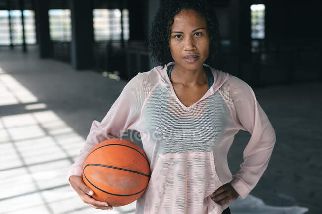 Portrait d'une femme afro-américaine debout dans un bâtiment urbain vide et tenant un ballon de basket. forme physique urbaine mode de vie sain. — Photo de stock