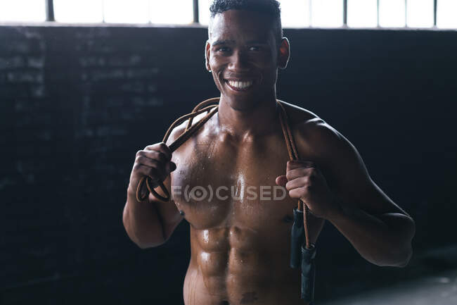 Ritratto di uomo afroamericano che salta la corda sulle spalle in un edificio urbano vuoto. guardando la telecamera e sorridendo. fitness urbano stile di vita sano. — Foto stock