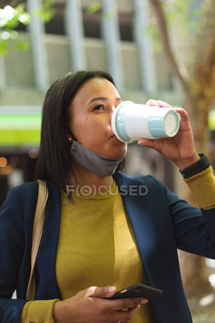 African american woman with lowered face mask drinking coffee on the street. lifestyle living during coronavirus covid 19 pandemic. — Stock Photo