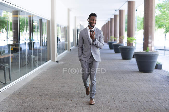 Hombre de negocios afroamericano caminando usando mascarilla hablando en smartphone. hombre de negocios en la salida en la ciudad. - foto de stock