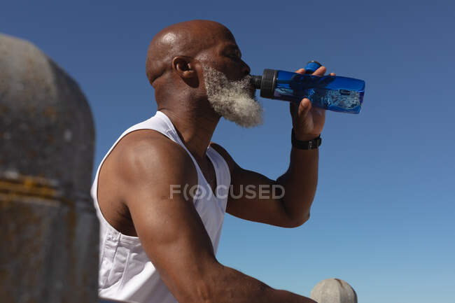 Ajuste o homem americano africano sênior que senta-se bebendo da garrafa de água contra o céu azul. tecnologia de aposentadoria saudável comunicação ao ar livre fitness lifestyle. — Fotografia de Stock