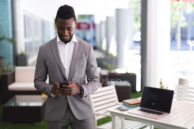 Hombre de negocios afroamericano de pie en un café y usando un teléfono inteligente. hombre de negocios en la salida en la ciudad. - foto de stock