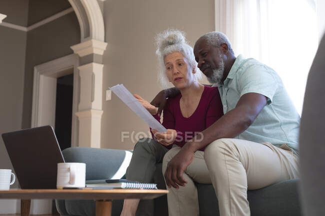 Senioren-Mischlingspaar mit Laptop im Wohnzimmer beim Bezahlen von Rechnungen. Während der Quarantäne zu Hause bleiben und sich selbst isolieren. — Stockfoto