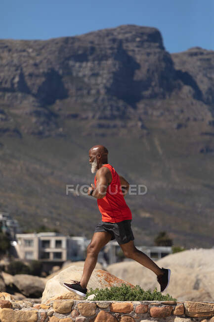 Fit senior african american man exercising running on rocky path beside mountain. healthy retirement outdoor fitness lifestyle. — Stock Photo