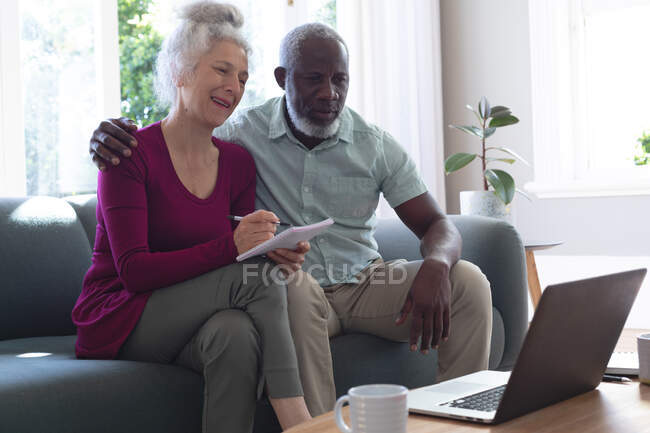 Pareja de raza mixta senior usando laptop pagando cuentas juntos en la sala de estar. permanecer en casa en aislamiento durante el bloqueo de cuarentena. - foto de stock
