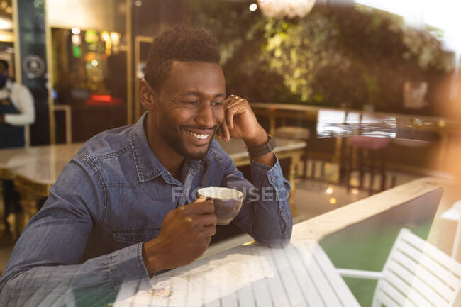 Hombre afroamericano sentado en un café bebiendo una taza de café y sonriendo. hombre de negocios en la salida en la ciudad. - foto de stock
