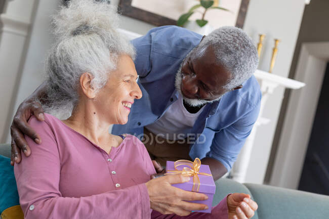 Старшая пара смешанных рас в гостиной мужчина дарит женщине подарок. оставаться дома в изоляции во время карантинной изоляции. — стоковое фото