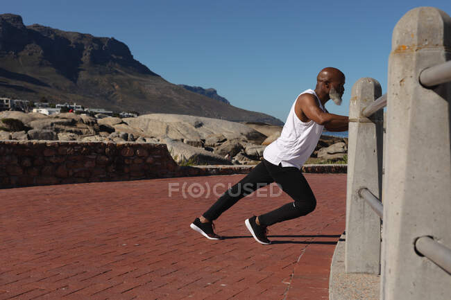 Ajuste hombre afroamericano senior ejerciendo apoyándose en estiramiento de fémur. retiro saludable estilo de vida fitness al aire libre. - foto de stock