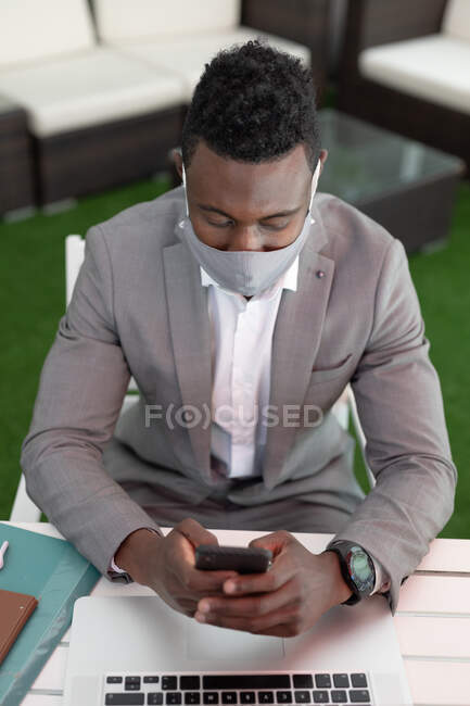 Afrikanisch-amerikanischer Geschäftsmann sitzt mit Gesichtsmaske in einem Café und benutzt Smartphone und Laptop. Geschäftsmann unterwegs in der Stadt. — Stockfoto